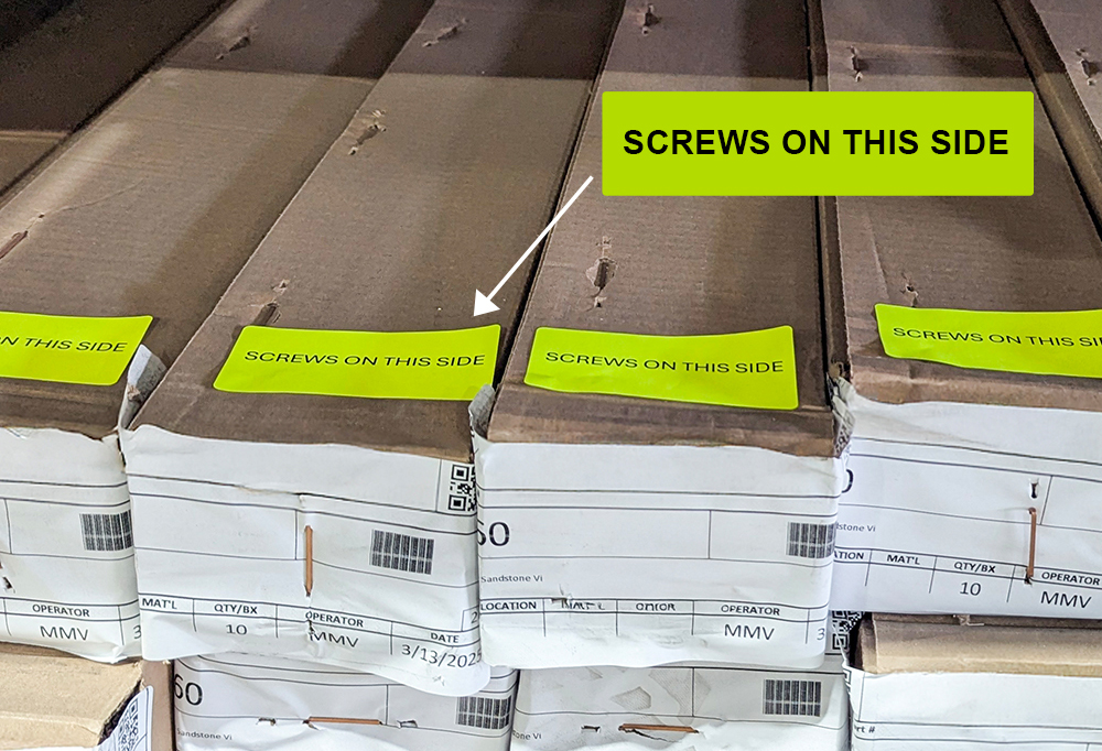 EDITED BOX Warehouse perspective of multiple cardboard boxes of 5001 weatherstripping stacked on a pallet. Bright, fluorescent yellow stickers with the text "SCREWS ON THIS SIDE" are placed on the end of each box to provide clear handling instructions.